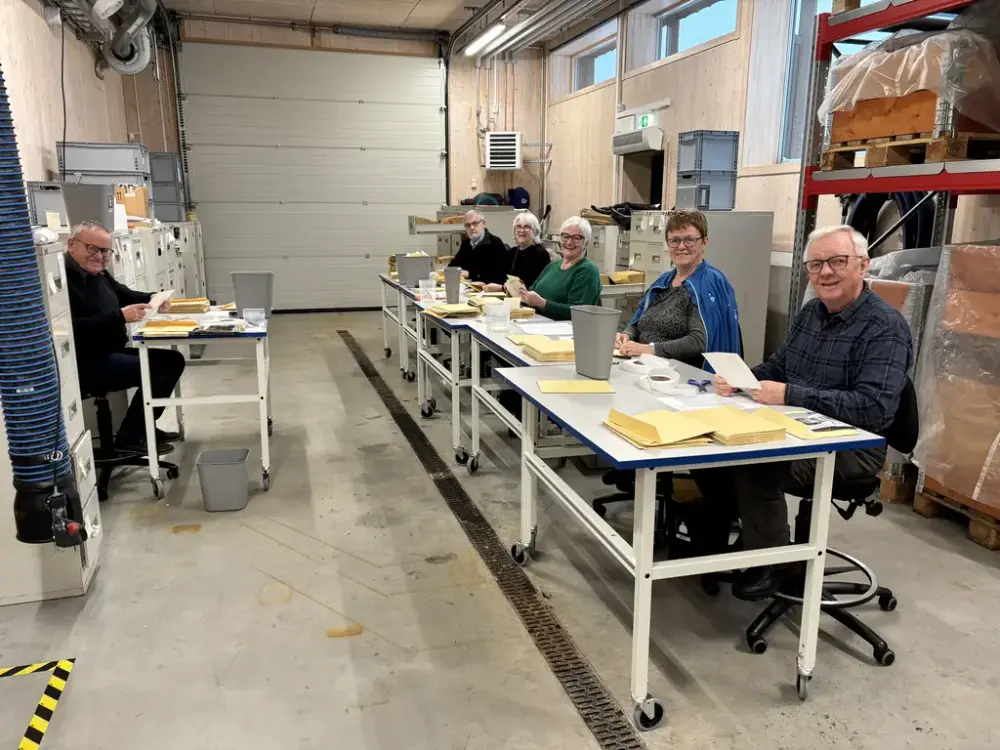 En gruppe eldre mennesker sitter ved et langbord i et lager og sorterer papirer