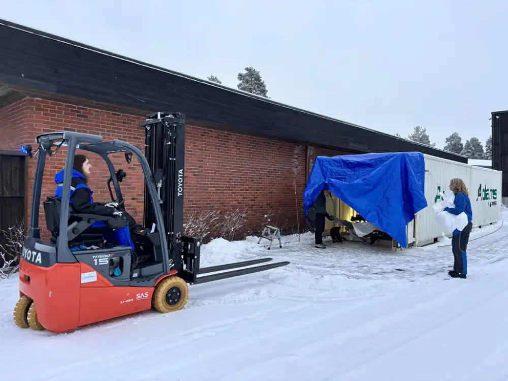 En person sitter i en gaffeltruck utendørs og en person står ved siden av