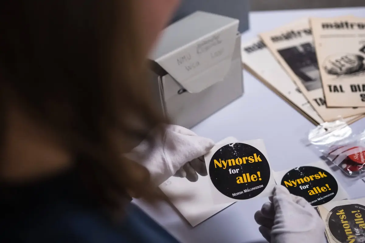 Person med kvite hanskar held eit svart klistremerke med teksten "Nynorsk for alle! NORSK MÅLUNGDOM" i gul og kvit skrift.