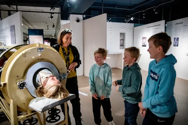 En omviser med 3 barn smiler og ser på en elev som ligger i jernlunga