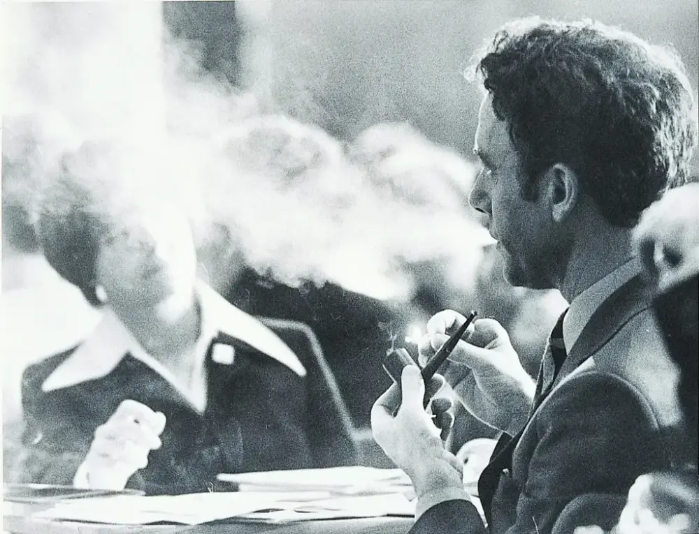 En mann på høyre side av bildet røyker pipe og røyklegger en kvinne til venstre i bildetø