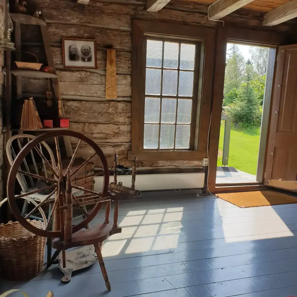 Døren står åpen til laftet hus. Solen skinner inn gjennom vindu og dør og kaster skygge på gulvet. En rokk og en stor står i ene kanten. På veggen henger et gammelt foto, i hjørne en hylle med forskjellige saker