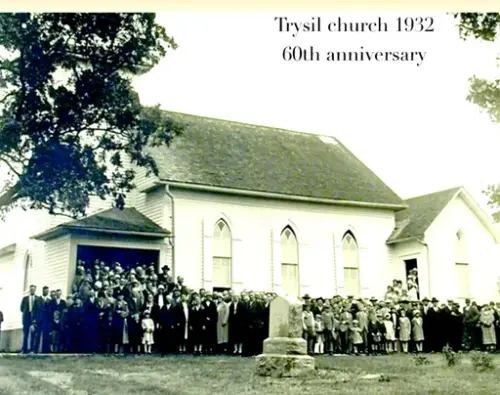 Trysil kirke fra siden med menigheten som står foran. Kirken er bygd i tre, og har tre høye gotiske/spisse vinduer på siden. 