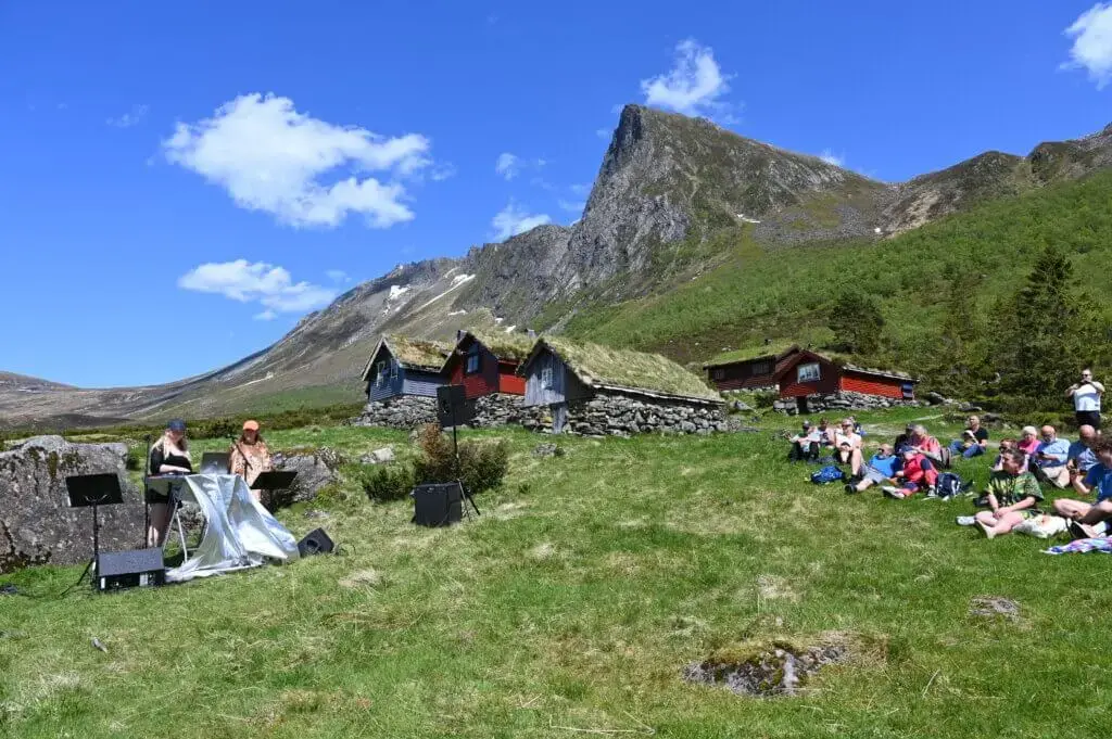 Utekonsert under blå, nesten skyfri himmel. Utøvarar og publikum står og sit på den grøne setervollen med seterhus og tindar som bakgrunn.