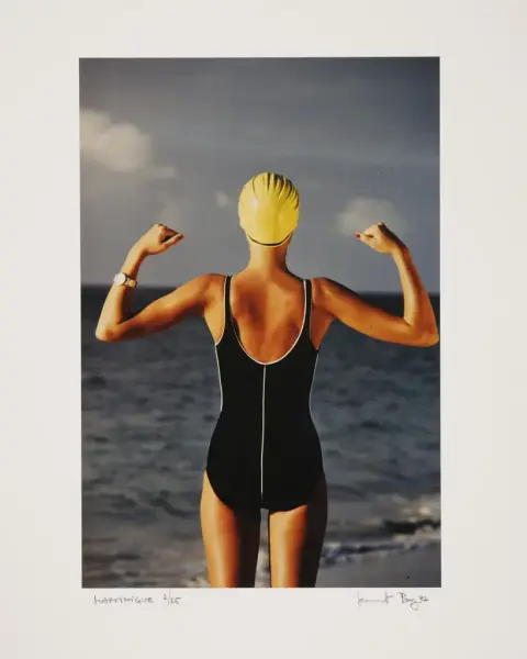 En kvinne står med ryggen til i svart badedrakt og gul badehette, på en strand der sola skinner. Hun knytter hendene og står i en slags sierspositur. 