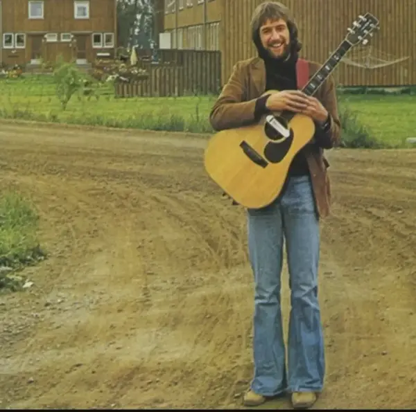 "Stein Ove Berg står smilende på en grusvei med en akustisk gitar i hendene. Han er iført en brun jakke, svart genser og slengbukser, med et landlig boligområde i bakgrunnen.