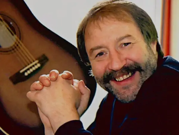 Stein Ove Berg smiler varmt mot kameraet med hendene foldet foran seg. I bakgrunnen henger en klassisk gitar.