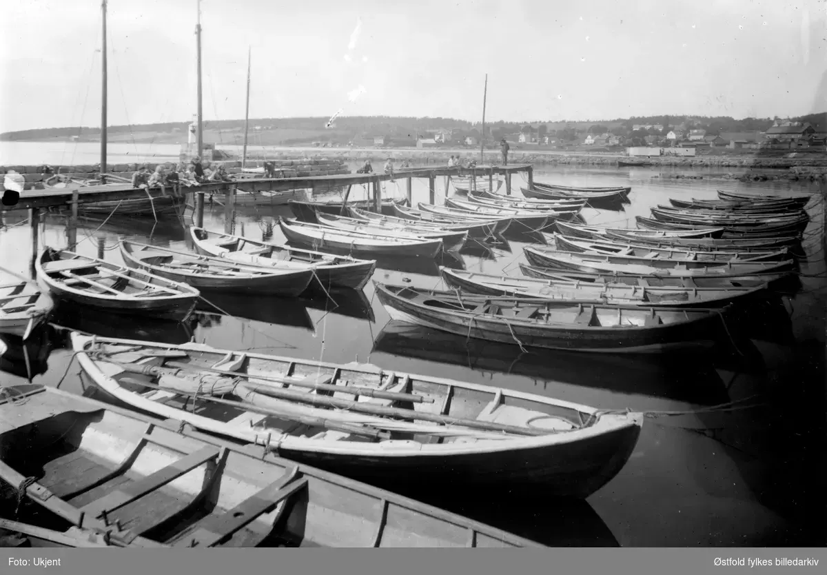 Sort-hvitt bilde med cirka 20 robåter og en brygge.