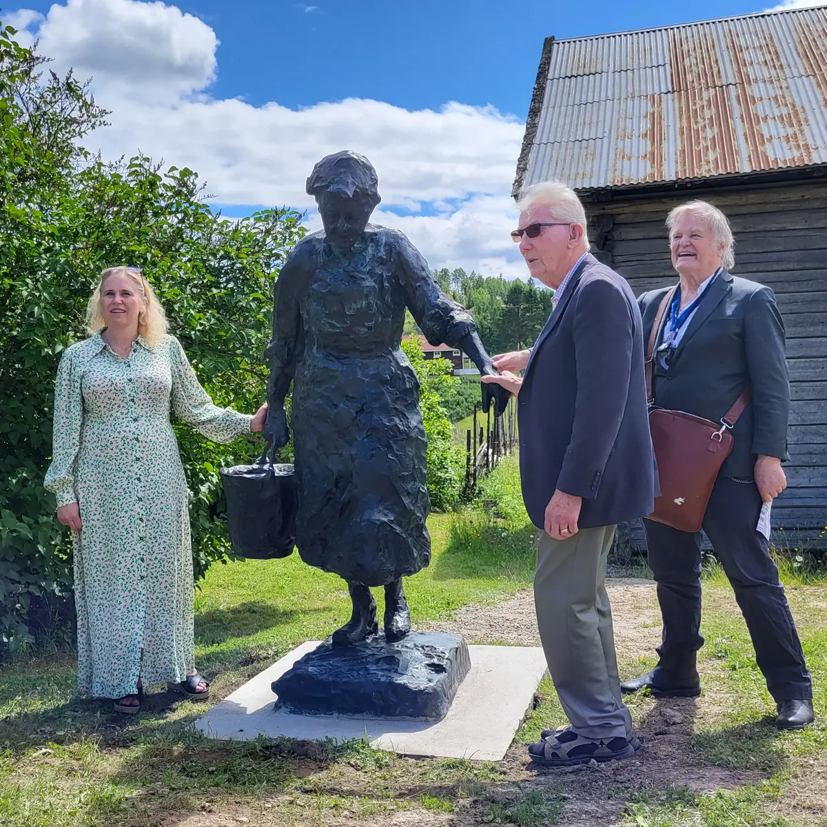 En kvinne og to eldre menn står ute i sommerværet, og holder i hendene på en statue av en kvinne som bærer en bøtte med vann