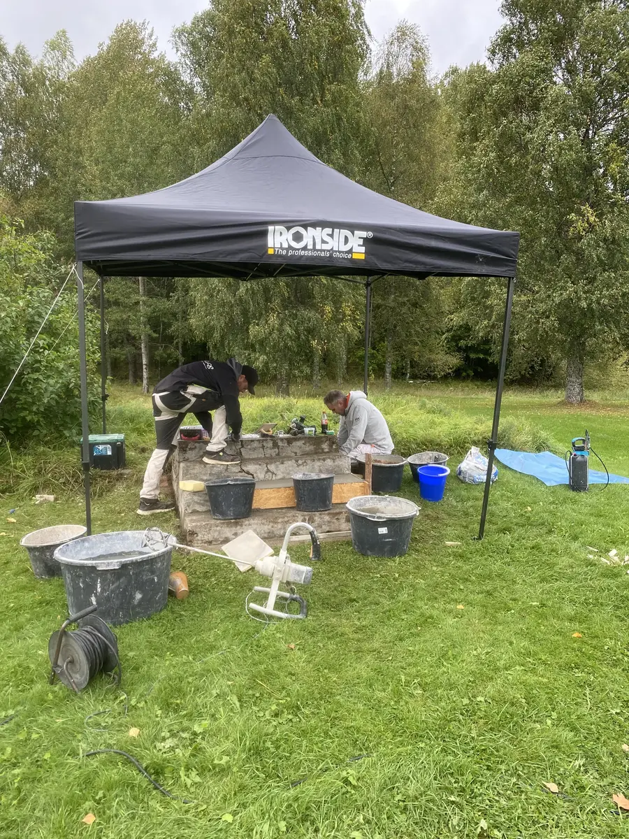 To menn står under et pop-up telt og jobber med en steintrapp som står ute i åpent naturlandskap. Rundt dem i gresset ligger diverse verktøy.