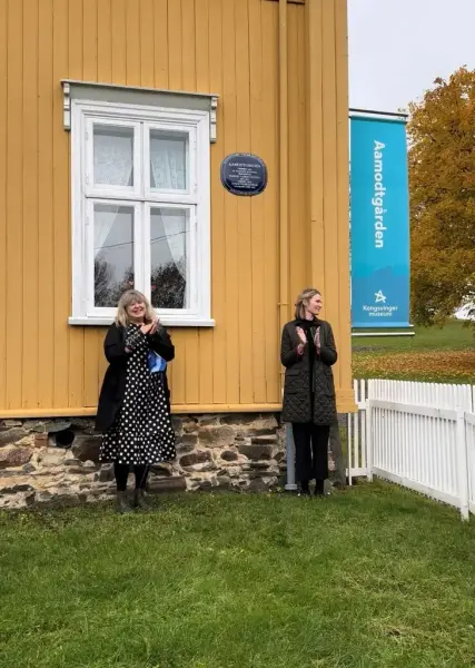 To kvinner står og klapper med hendene ved en gul husvegg, med et blått rundt skilt. 