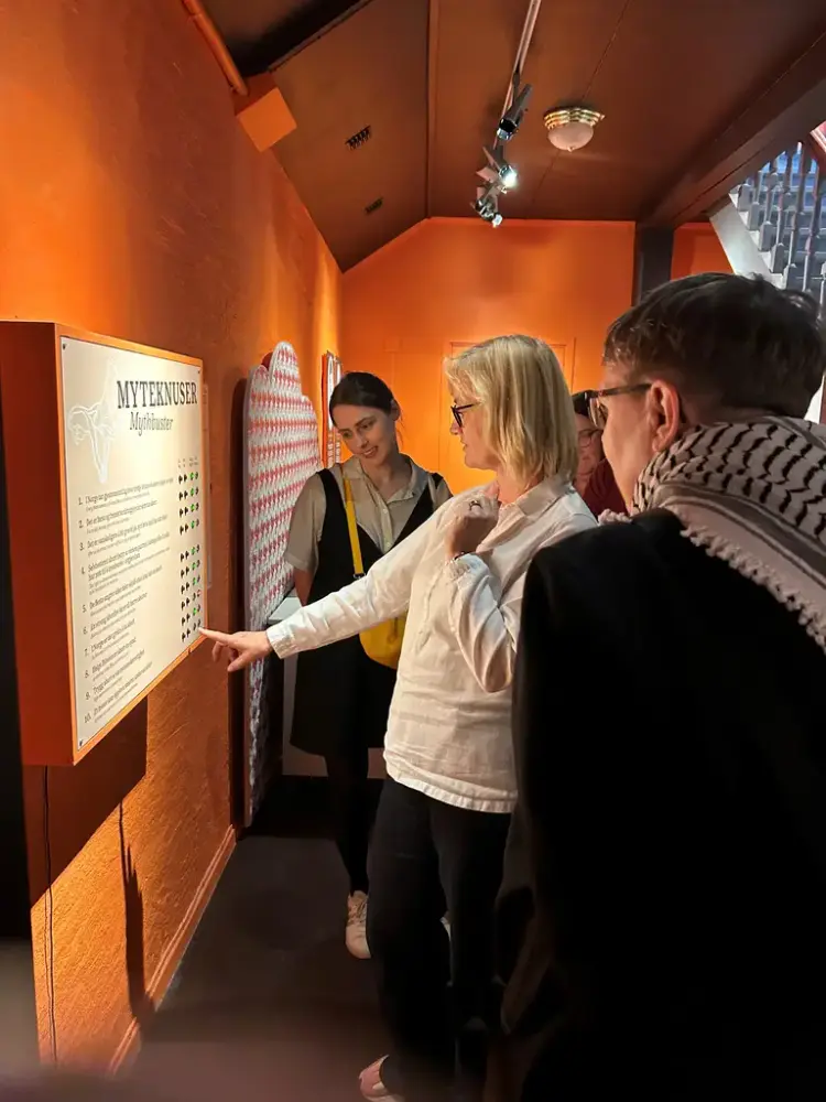 En gruppe mennesker står samlet rundt et skilt som henger på en oransje vegg, og en kvinne i front peker på nedre del av skiltet. 