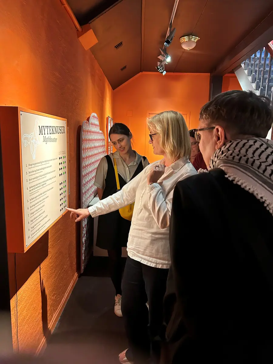En gruppe mennesker står samlet rundt et skilt som henger på en oransje vegg, og en kvinne i front peker på nedre del av skiltet. 