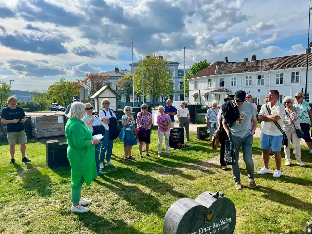 En stor gruppe mennesker står samlet ute på en gravlund, og ser på en kvinne iført grønne klær snakke. 