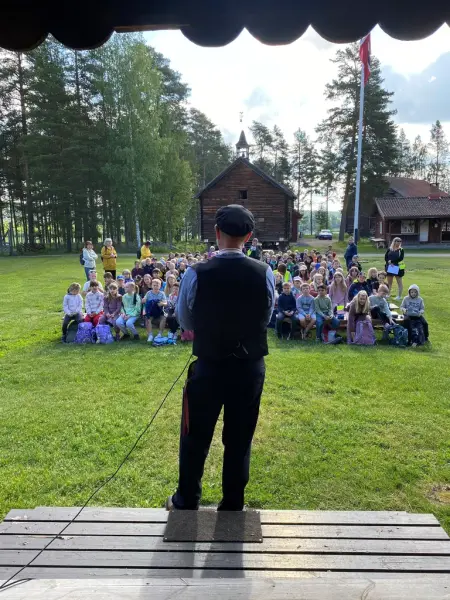 En mann iført klær fra gamledager, taler til en stor gruppe med unge elever som sitter foran han på gresset.