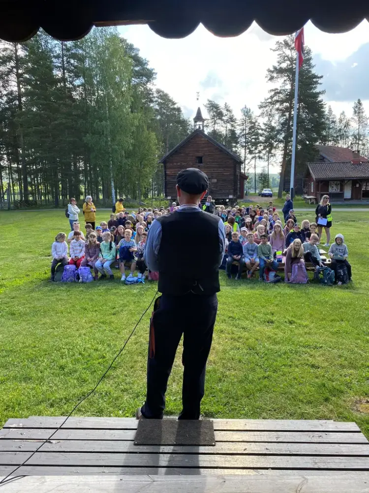 En mann iført klær fra gamledager, taler til en stor gruppe med unge elever som sitter foran han på gresset.