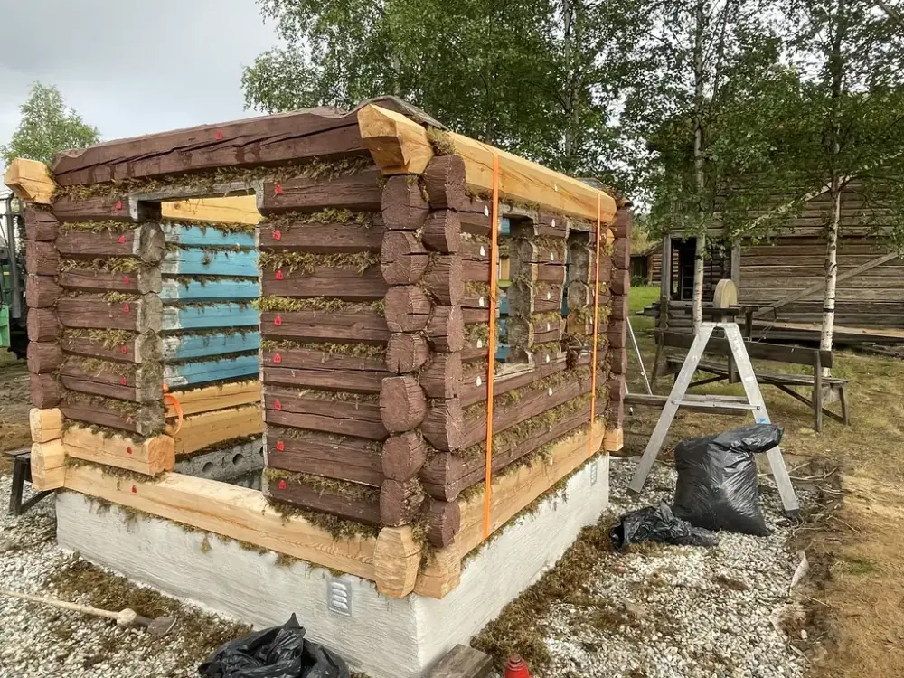 Et lekehus av laftet tømmer som står uten tak, vinduer og dør.