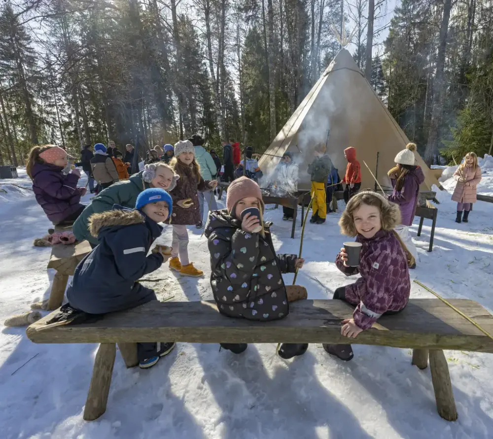 Barn sitter på benk i snøen og drikker varm drikke ved bålplass utendørs.