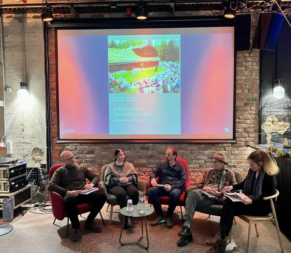 En gruppe personer sitter i hver sin røde stol i en halvsirkel, foran en stor skjerm som viser forsiden av en bok