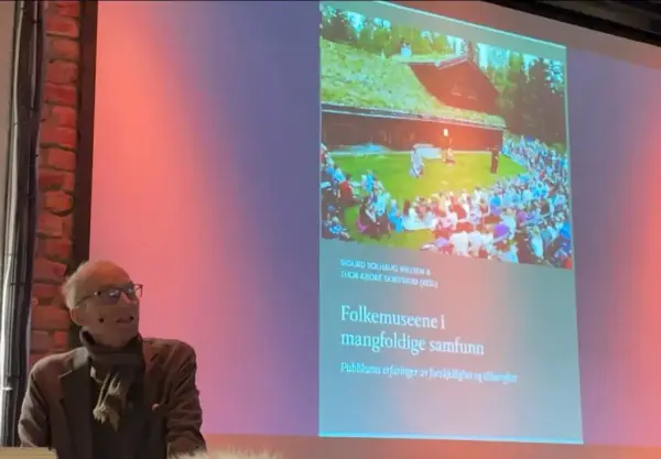 En eldre mann sitter og snakker foran en skjerm som viser forsiden av en bok