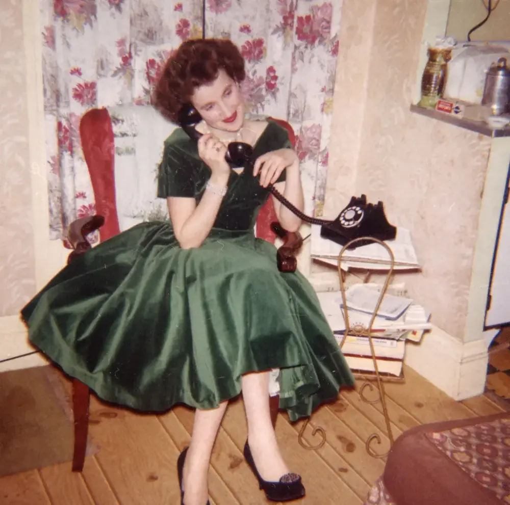 Kvinne i grønn kjole sitter på en rød stol og snakker i en sort telefon. Blomstrete gardiner i bakgrunn.