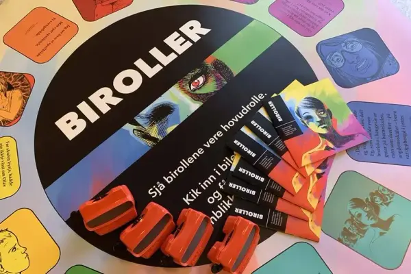 Del av utstillinga. Duk med fargerike illustrasjonar, oransje biletkikkertar og ein bunke hefte.