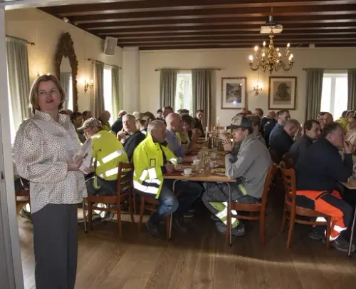 Menn i arbeidsklær sitter til bords under kranselag, en kvinne holder tale.