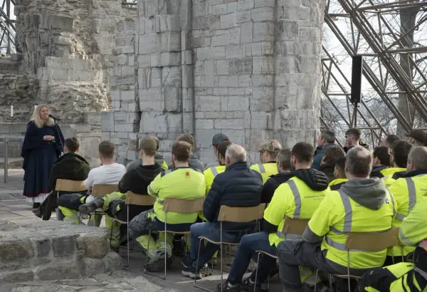 Menn i arbeidsklær sitter og hører på kvinne i middelalderklær som synger gregoriansk sang i domkirkeruinen.