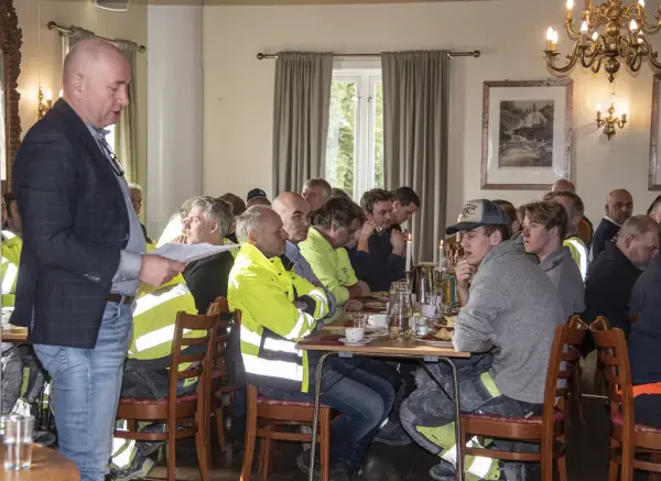 Menn i arbeidsklær sitter til bords under kranselag, Annos direktør holder tale.
