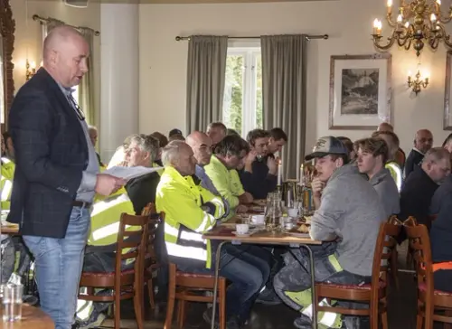 Menn i arbeidsklær sitter til bords under kranselag, Annos direktør holder tale.