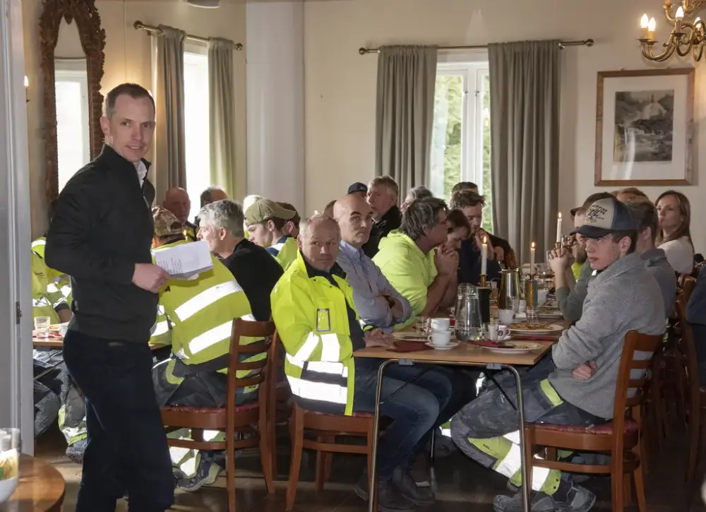 Menn i arbeidsklær sitter til bords under kranselag, en mann holder tale.