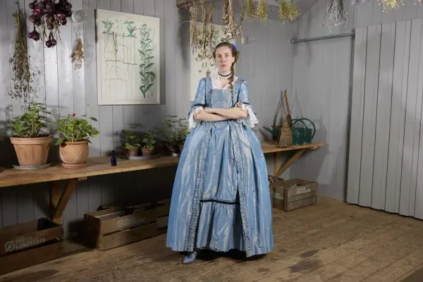 Brigitte Le Bleu, en karakter fra påskekrimmen 2025 på Norsk Folkemuseum, kledd i en blå kjole, står omgitt av tørkede urter og potteplanter.