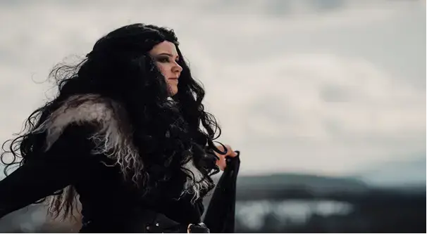 Fotografi av en ung kvinne med langt, tykt svart hår i profil. Hun har armene ut til siden og er kledd i svarte klær og en grå pels over skuldrene. Hun har svart sminke rundt øynene og smiler. I bakgrunnen er et grått lamdskap med mørk himmel , trør og vann.