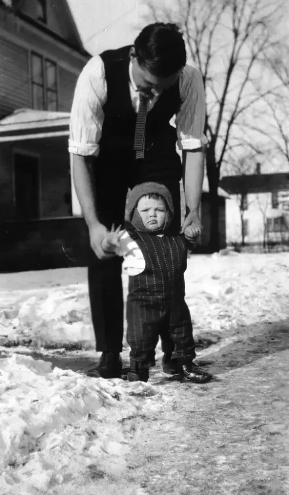 En mann holder armene til et lite barn som står. Snø på bakken. Bygninger bak.