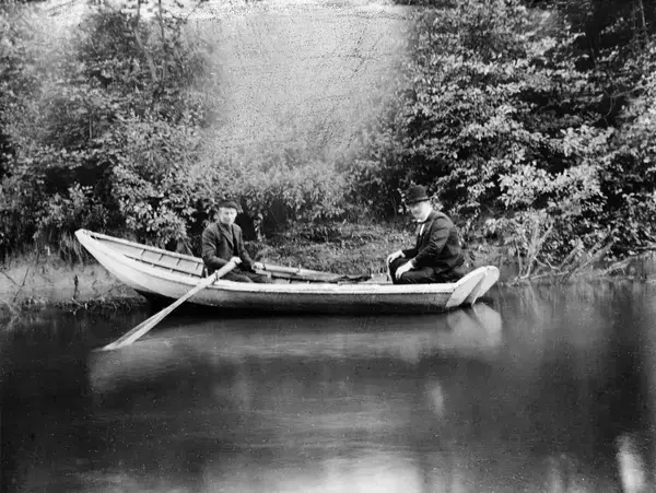 Bilde fra 1903, tatt av en ukjent fotograf, viser to menn i en båt på Alnaelvens nedre løp. De sitter rolig i båten, omgitt av frodig vegetasjon og stille vann.