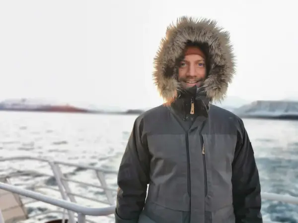 dreas Østhagen, seniorforsker ved Fridtjof Nansens Institutt i Oslo, står smilende på et fartøy iført vinterjakke med pelshette. Bakgrunnen viser snødekte fjell og et rolig hav.