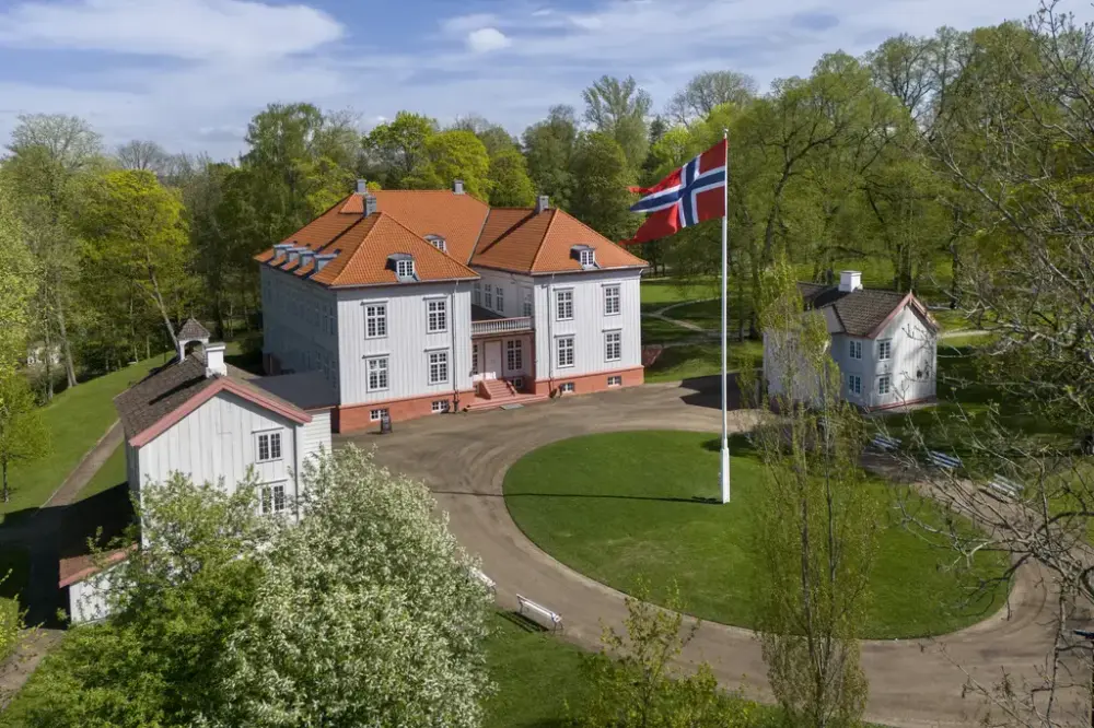 Eidsvollsbygningen sett fra luften. Bygningen står omgitt av grønt landskap, med det norske flagget som vaier foran inngangen