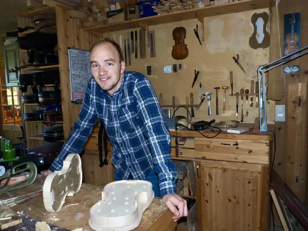 En mann i blå, rutete skjorte står i et verksted med en uferdig fele foran seg.