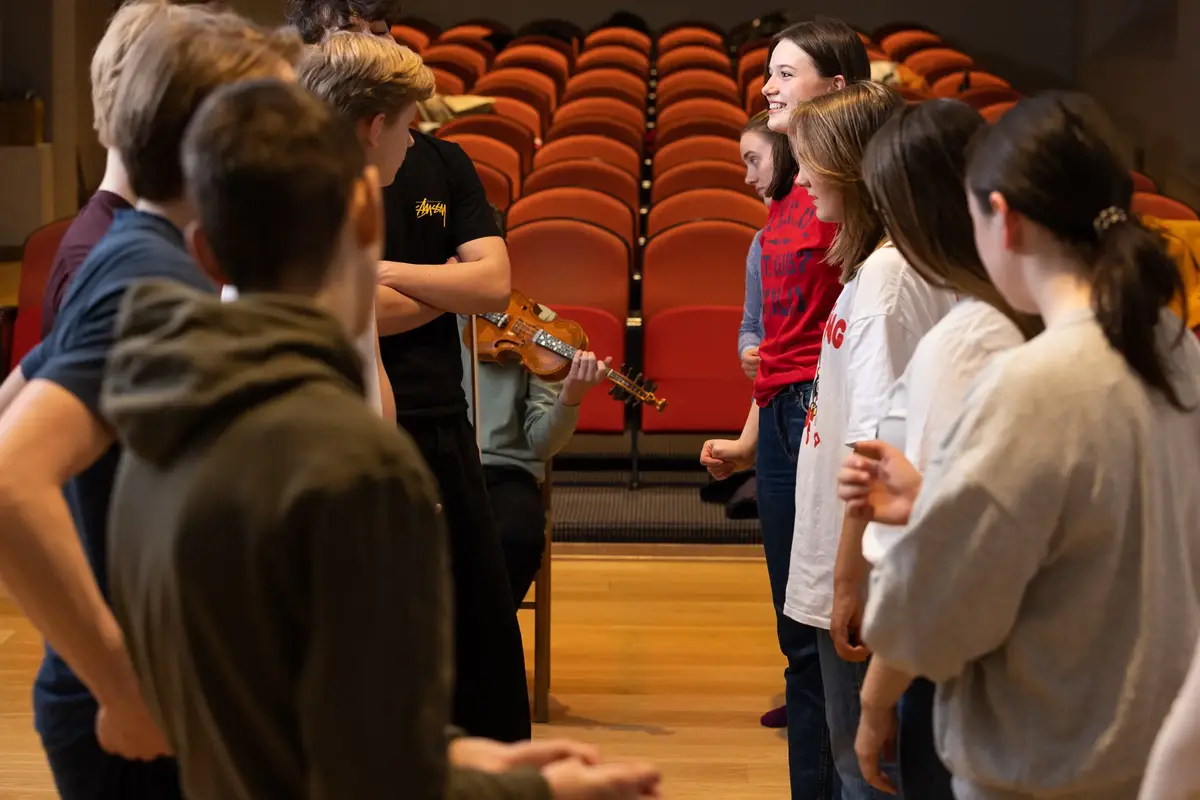 Ungdommer øver på norsk folkedans med felespiller i bakgrunnen, i en sal med røde stoler.