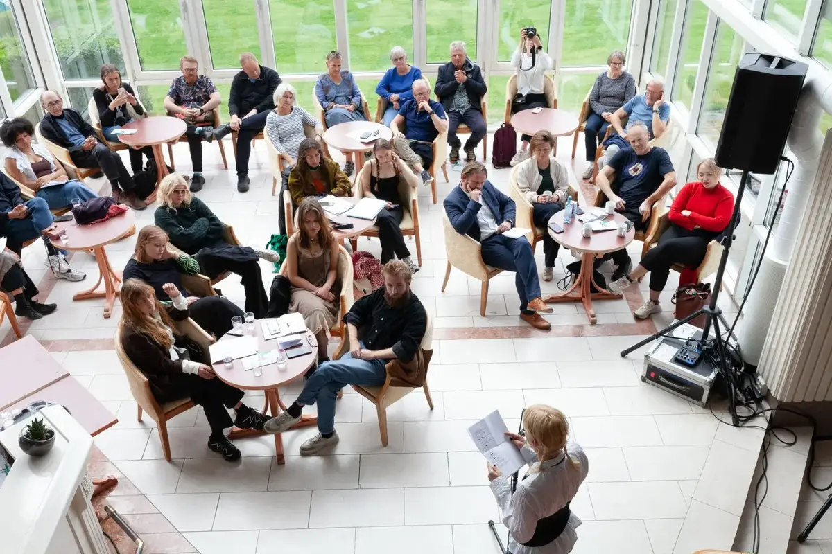 nokre titals personar sit rundt runde bord i eit utstikk og høyrer på nokon som står og les