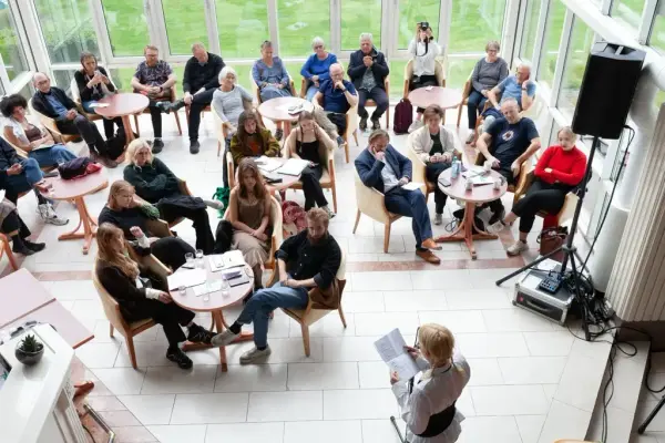 nokre titals personar sit rundt runde bord i eit utstikk og høyrer på nokon som står og les