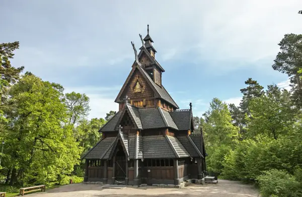 Stavkirken fra Gol, omgitt av grønt løvverk, fotografert på Norsk Folkemuseum en klar sommerdag.