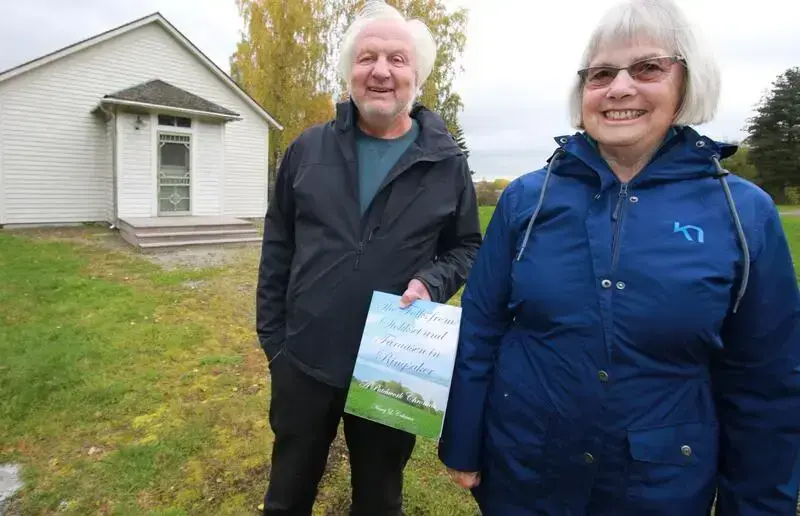 En mann og en kvinne med grått hår og allværsjakker står foran et hvit hus. Mannen viser frem en bok.