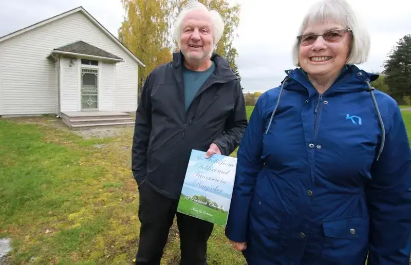 En mann og en kvinne med grått hår og allværsjakker står foran et hvit hus. Mannen viser frem en bok.
