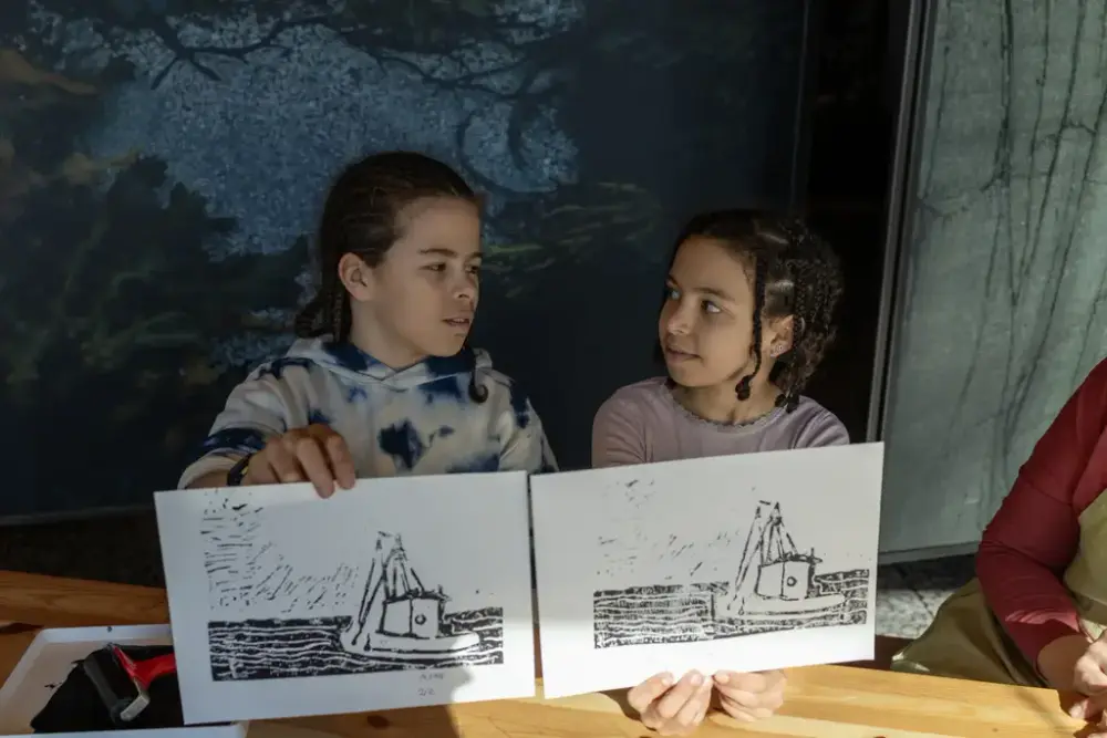 To barn holder opp hver sin grafiske trykk av en båt, og ser på hverandre ved bordet i grafikkverkstedet.