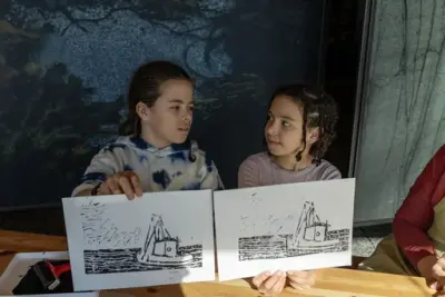 To barn holder opp hver sin grafiske trykk av en båt, og ser på hverandre ved bordet i grafikkverkstedet.