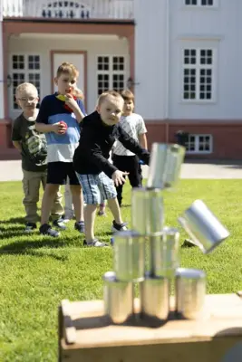 Barn deltar i blikkbokskasting med erteposer på plenen foran Eidsvollsbygningen, som del av uteaktiviteter på museet.