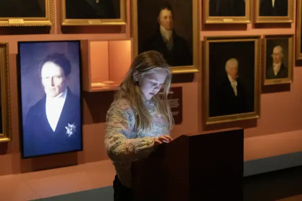 Jente utforsker en interaktiv skjerm i utstillingen i demokratisenteret Wergelands Hus på Eidsvoll 1814, omgitt av portretter av eidsvollsmennene.