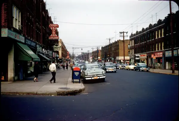 Streetscape from 'Lapskaus Boulevard' in New York, with bustling shops, passersby, and vintage cars.