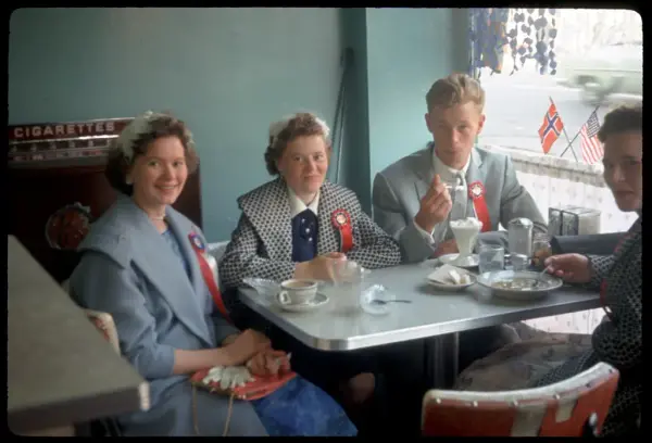 Café-goers on Lapskaus Boulevard, adorned with May 17th ribbons, with Norwegian and American flags in the window.